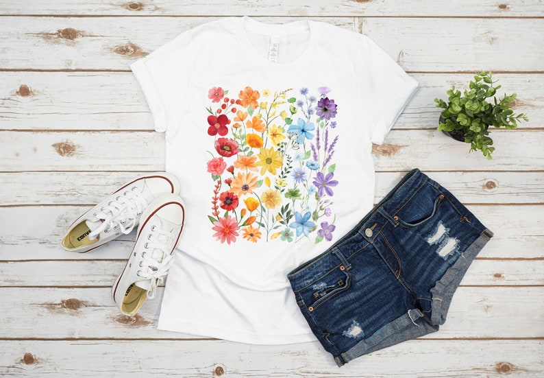 Puede incluir: Una camiseta blanca con un dise&ntilde;o floral arco&iacute;ris. Las flores est&aacute;n pintadas en acuarela y dispuestas en una franja vertical. La camiseta se combina con pantalones cortos de mezclilla y zapatillas blancas.