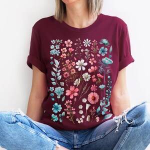 May include: A maroon t-shirt with a floral print featuring pink, blue, and white flowers. The flowers are arranged in a vertical pattern on the front of the shirt.