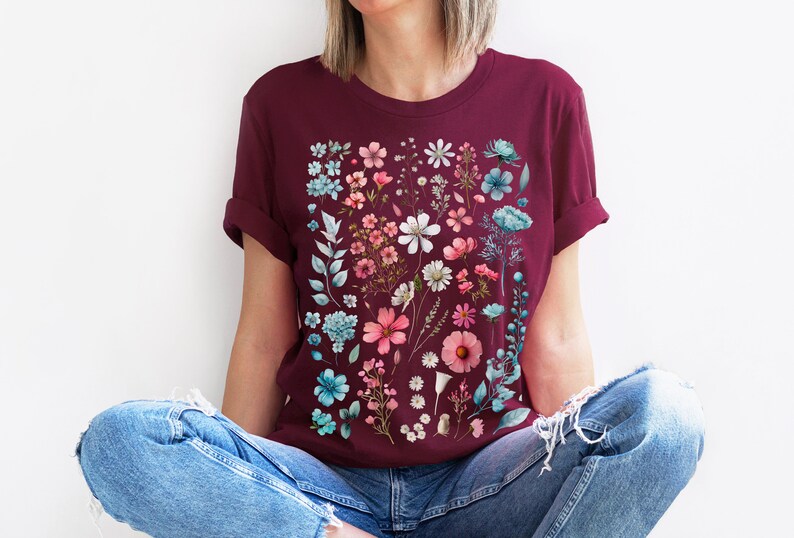 May include: A maroon t-shirt with a floral print featuring pink, blue, and white flowers. The flowers are arranged in a vertical pattern on the front of the shirt.