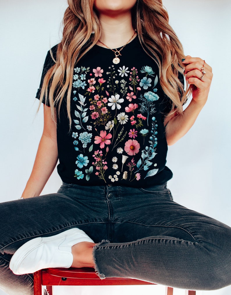 May include: A black t-shirt with a floral print featuring a variety of pink, blue, and white flowers. The flowers are arranged in a vertical pattern on the front of the shirt.