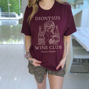 May include: A maroon t-shirt with a white graphic of a person holding a glass of wine and two bottles of wine. The text "Dionysus Wine Club Charter Member" is printed on the shirt.