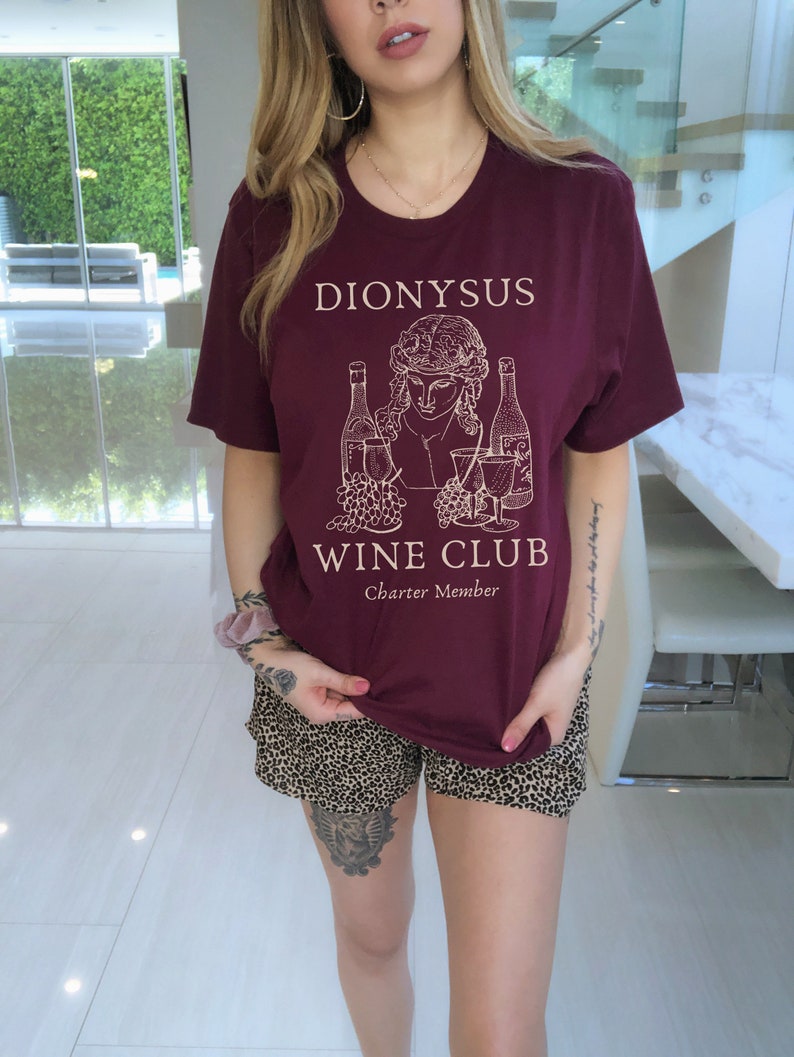 May include: A maroon t-shirt with a white graphic of a person holding a glass of wine and two bottles of wine. The text "Dionysus Wine Club Charter Member" is printed on the shirt.