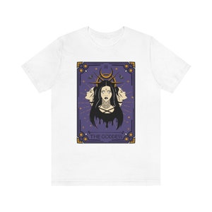 May include: White t-shirt with a purple and gold tarot card design. The card depicts a woman with three faces and the text "THE GODDESS" at the bottom.