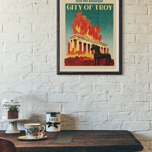 May include: A vintage travel poster with a dark wood frame. The poster features a blue background with the text "visit the beautiful CITY OF TROY" in white. The poster depicts a burning building with a black horse standing in front of it.