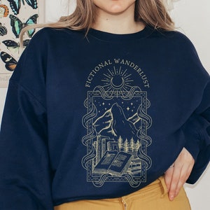 Op de afbeelding: Een donkerblauw sweatshirt met een grafisch ontwerp van een bergketen, een boek en een halve maan. De tekst "Fictional Wanderlust" is gedrukt in een gouden schrijfletter.