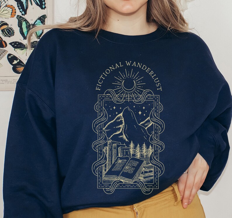 Op de afbeelding: Een donkerblauw sweatshirt met een grafisch ontwerp van een bergketen, een boek en een halve maan. De tekst "Fictional Wanderlust" is gedrukt in een gouden schrijfletter.