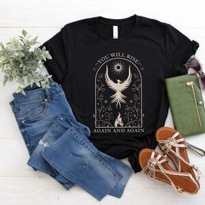 May include: Black t-shirt with a graphic of a phoenix rising from flames in a circle with the text "You Will Rise Again and Again".