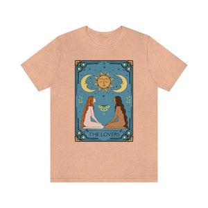 May include: A peach-colored t-shirt with a graphic of the Lovers tarot card. The card features two figures, a sun, a moon, and a moth. The text "THE LOVERS" is printed below the figures.