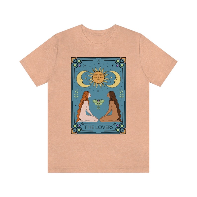May include: A peach-colored t-shirt with a graphic of the Lovers tarot card. The card features two figures, a sun, a moon, and a moth. The text "THE LOVERS" is printed below the figures.