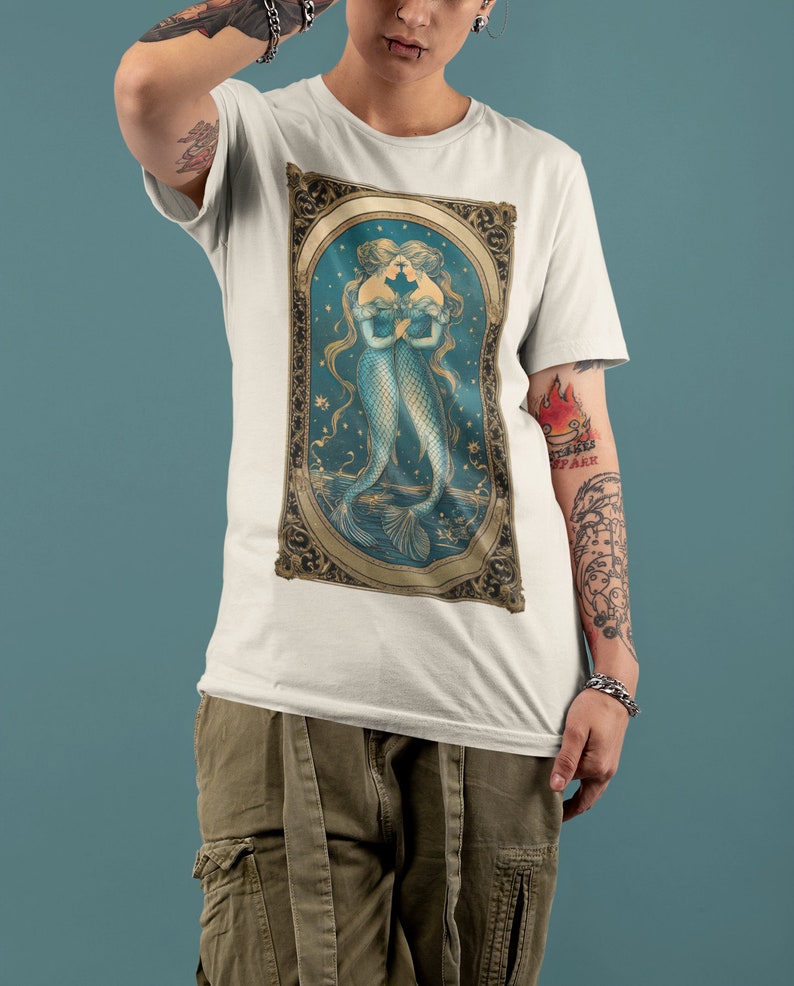 Puede incluir: Camiseta blanca con una ilustraci&oacute;n vintage de dos sirenas con las colas entrelazadas, enmarcada en oro sobre un fondo azul y dorado.