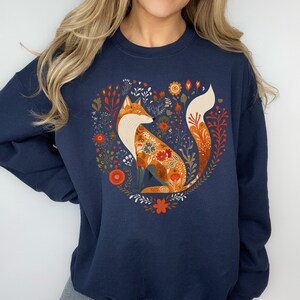 May include: A navy blue sweatshirt featuring a graphic of a fox surrounded by flowers in a heart shape. The fox is orange and white with floral details.
