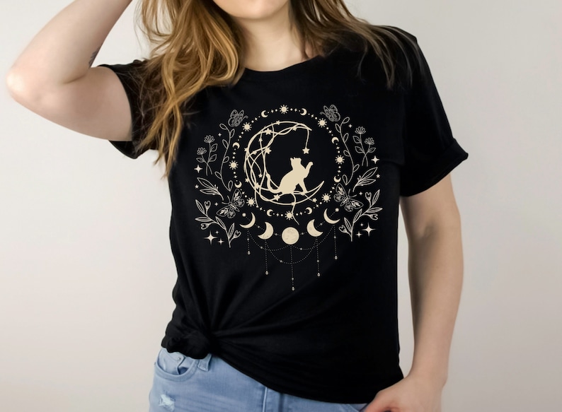 May include: Black t-shirt with a white graphic of a cat sitting on a crescent moon surrounded by stars, flowers, and moons.