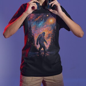 May include: A black t-shirt with a colorful graphic print of a Bigfoot walking through a forest under a starry sky.