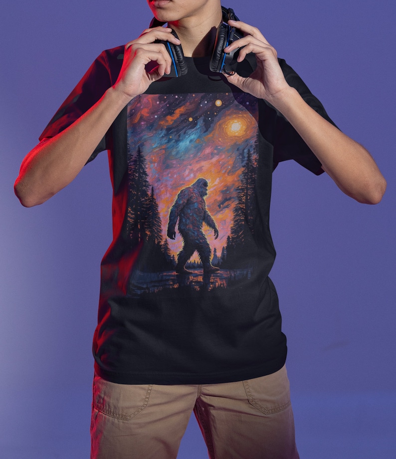 May include: A black t-shirt with a colorful graphic print of a Bigfoot walking through a forest under a starry sky.