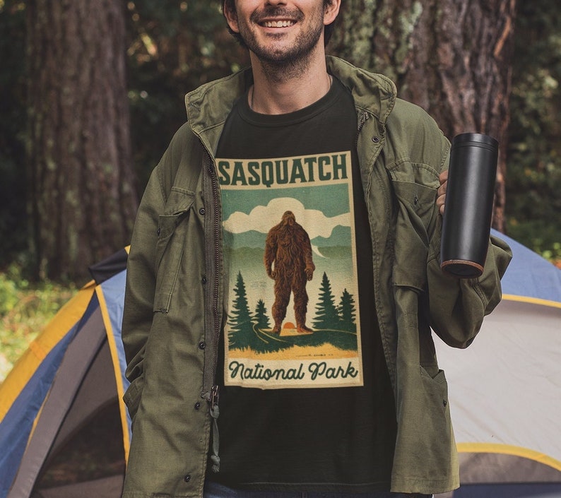 May include: Black t-shirt featuring a vintage-style graphic of a Sasquatch in a National Park setting. The design includes the text "SASQUATCH" at the top and "National Park" at the bottom, with a large Bigfoot illustration in the center.