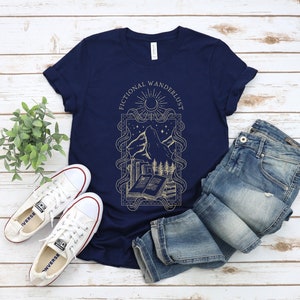 K&ouml;nnte beinhalten: Ein marineblaues T-Shirt mit einem goldenen Grafikdesign, das einen Berg, ein Buch und den Text "Fictional Wandererlust" zeigt.