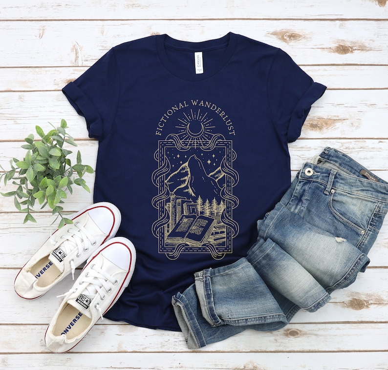 K&ouml;nnte beinhalten: Ein marineblaues T-Shirt mit einem goldenen Grafikdesign, das einen Berg, ein Buch und den Text "Fictional Wandererlust" zeigt.