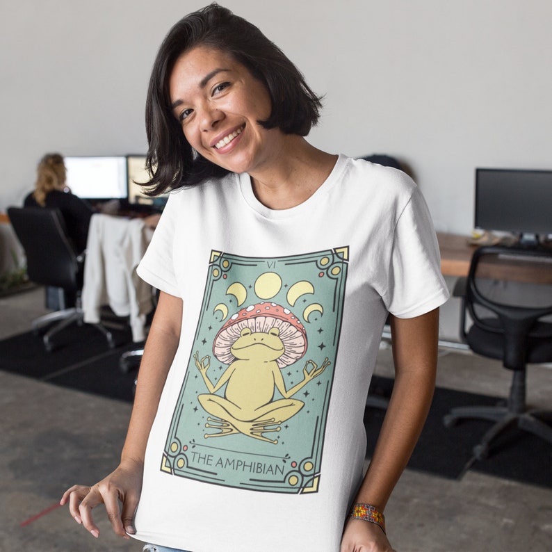May include: White t-shirt with a graphic of a green frog sitting in a red and white mushroom with a yellow moon and stars on a green background. The text "THE AMPHIBIAN" is below the frog.