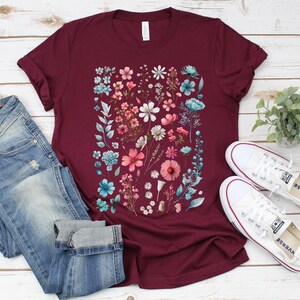 May include: A maroon t-shirt with a floral print featuring pink, blue, and white flowers. The shirt is paired with blue jeans and white sneakers.