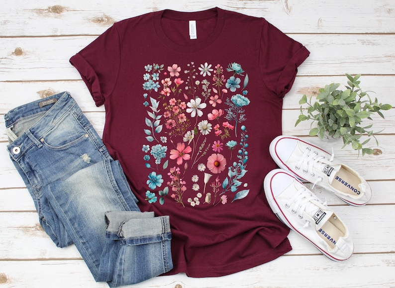 May include: A maroon t-shirt with a floral print featuring pink, blue, and white flowers. The shirt is paired with blue jeans and white sneakers.