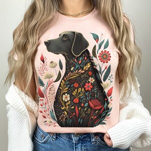 May include: A pink t-shirt with a black dog silhouette surrounded by colorful flowers. The dog is looking to the left and has a floral pattern on its body.