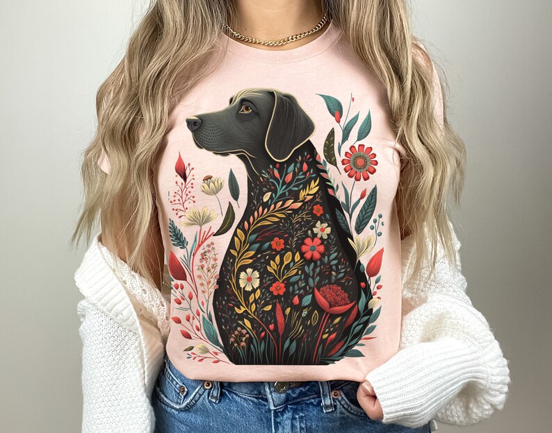 May include: A pink t-shirt with a black dog silhouette surrounded by colorful flowers. The dog is looking to the left and has a floral pattern on its body.