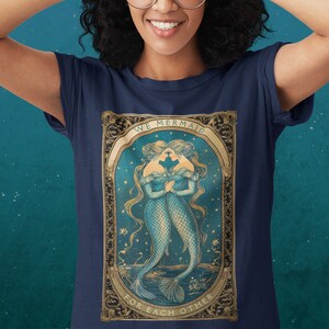 Op de afbeelding: Een marineblauw t-shirt met een vintage illustratie van twee zeemeerminnen met de tekst "We Mermaid For Each Other" in een gouden frame.