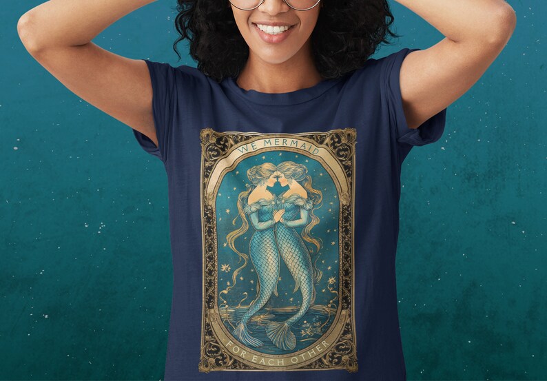 Op de afbeelding: Een marineblauw t-shirt met een vintage illustratie van twee zeemeerminnen met de tekst "We Mermaid For Each Other" in een gouden frame.