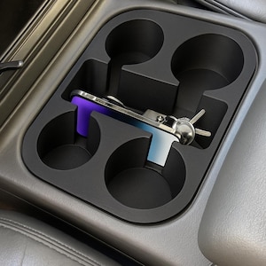 May include: A black car console cup holder with four circular compartments. A silver and purple phone is placed in one of the compartments. A set of keys is also visible.