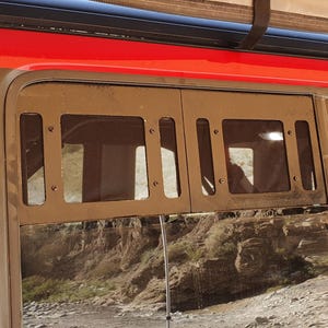 May include: A section of a red vehicle with a black roof rack and a tan awning. The side window has a metal protective cover with rectangular cutouts. The background shows a mountainous, arid landscape.