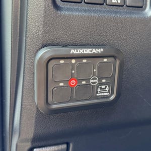 May include: Black control panel with the text "AUXBEAM" and multiple illuminated buttons. The panel includes buttons for windshield wipers, power outlets, and other vehicle functions. The power button is illuminated red.