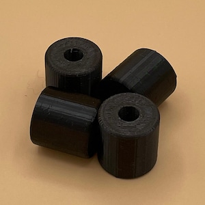 May include: Four black cylindrical auto parts with a hole through the center are arranged on a tan surface. The parts are made of a textured material and are likely 3D-printed. The Lazarus Auto Parts logo is visible in the upper right corner.