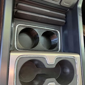 May include: A car's center console with two cup holders and a storage area. The cup holders are black with a textured surface and a silver trim. The console is a mix of black and tan colors.