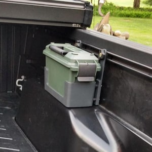 May include: A green military-style ammo box mounted inside a black truck bed. The box is rectangular with a hinged lid and a gray base. The truck bed has a black rubber mat and a red tailgate.