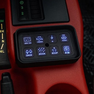 May include: Interior view of a vehicle's console, featuring a red dashboard with black controls. Includes a gear shift, transfer case selector, and a panel of illuminated switches labeled "DACK", "ROCK", "WHIP", and "AIR".