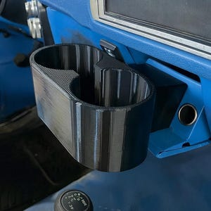 May include: A black, dual-cup holder, 3D-printed, mounted on a blue dashboard. The cup holder is positioned above a gear shift lever. The interior of the vehicle is blue.