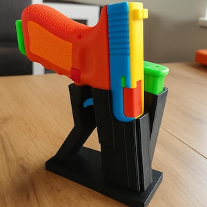 May include: A colorful toy gun with a textured orange grip, a blue slide, and a yellow top. The toy is held in a black stand. The magazine is green. The toy gun is designed with bright colors.