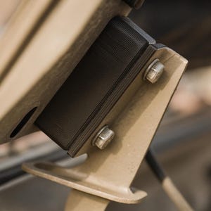 May include: Close-up of a black, rectangular component with a textured surface, secured to a beige metal bracket with two silver bolts. The background is blurred, suggesting an automotive or mechanical context.
