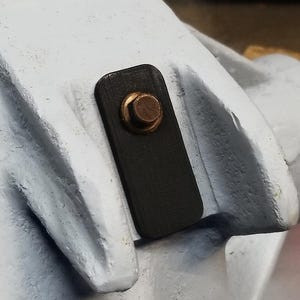 May include: Close-up of a black rectangular metal plate with a copper-colored rivet. The plate is attached to a white, textured metal surface. The image is well-lit, highlighting the details of the metalwork.