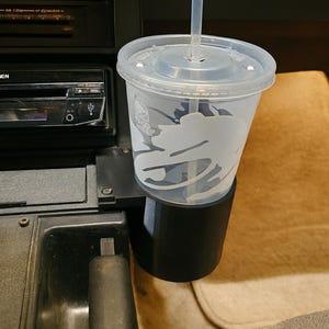 May include: A clear plastic cup with a lid and straw, featuring a white logo, sits in a black cup holder inside a vehicle. The cup holder is mounted on the dashboard near the radio.