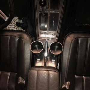 May include: Interior view of a classic car's front seats. The image shows the center console with two cup holders, a gear shift, and a leather armrest. The seats are dark brown leather, and the steering wheel is visible.