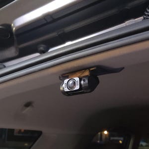 May include: A black and silver dash camera mounted inside a vehicle. The camera has a lens and several small lights. It is attached to the vehicle's ceiling.