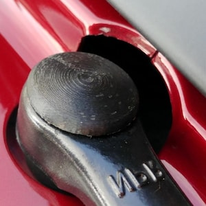 May include: Close-up of a black, textured knob with a circular pattern, set against a glossy red surface. The knob is part of a larger, dark metal component, with the letters "MB" visible.