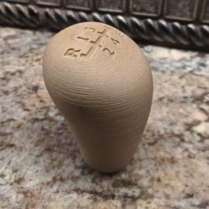 May include: A tan-colored shift knob with a gear pattern etched on top. The knob has a smooth, rounded shape and is designed for a vehicle's manual transmission. The background is a speckled countertop.