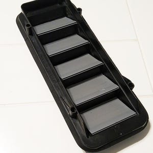 May include: A black plastic car vent with six angled slats. The vent has a rectangular shape with rounded corners and small mounting holes. The slats are a matte black, and the frame has a slightly textured surface.