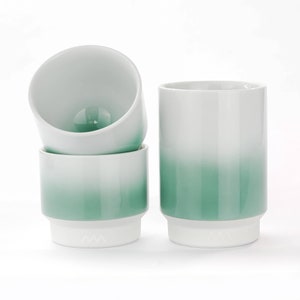 May include: Four white ceramic cups with a gradient green design. The cups have a cylindrical shape with a white base and a subtle logo. The cups are stacked and arranged on a white surface.