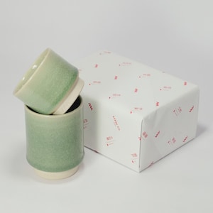 Op de afbeelding: Twee groene keramische bekers met een witte basis zijn naast een verpakte geschenkdoos gestapeld. De bekers hebben een getextureerde afwerking en een groen verloop. De geschenkdoos is verpakt in wit papier met rode golfpatronen en tekst.