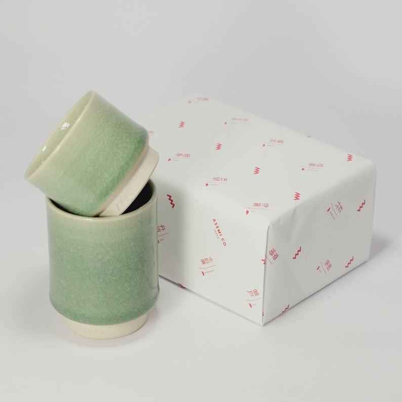 Op de afbeelding: Twee groene keramische bekers met een witte basis zijn naast een verpakte geschenkdoos gestapeld. De bekers hebben een getextureerde afwerking en een groen verloop. De geschenkdoos is verpakt in wit papier met rode golfpatronen en tekst.