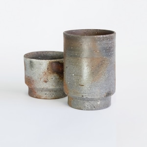 May include: Two handmade ceramic cups. The larger cup is cylindrical, and the smaller cup is a low bowl. Both have a speckled, textured surface with shades of gray, brown, and tan. The cups have a rustic, artisanal appearance.