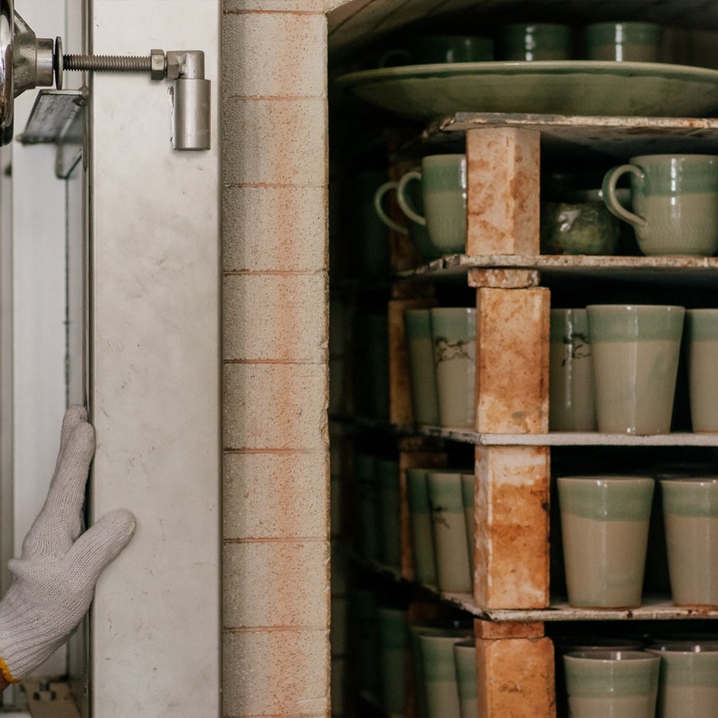 Op de afbeelding: Een oveninterieur vol met planken met keramische mokken en borden. De mokken zijn lichtgroen met een donkergroene rand. De ovendeur staat open en onthult de keramische stukken die klaar zijn om te bakken. Een gehandschoende hand is zichtbaar.